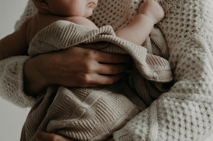 Mother wearing a cream cardigan holding a baby to her chest in a cream blanket